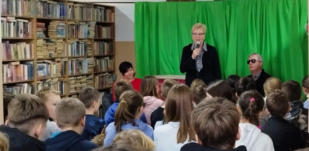 Zdjęcie ze spotkania, w centrum obrazu Alina Galas z mikrofonem, po prawej stronie Krzysztof Galas, z przodu głowy dzieci, po lewej stronie szefowa biblioteki.