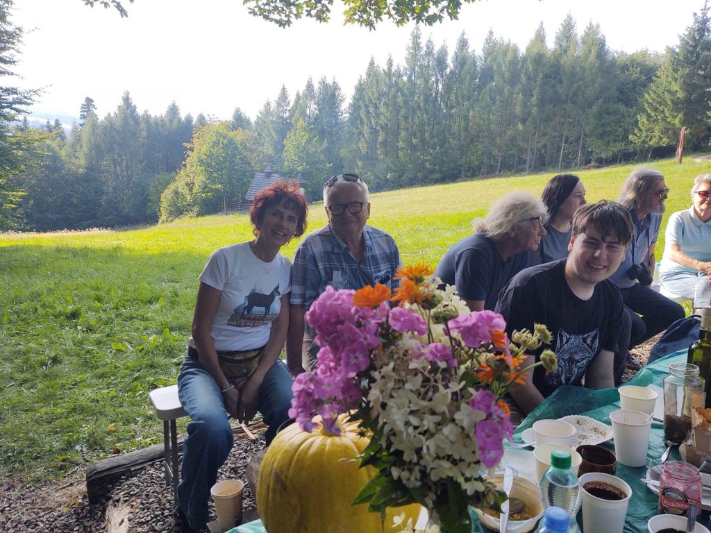 Na pierwszym planie bukiet kwiatów i dynia, za nimi grupa ludzi siedzących na ławce, w tle łąka, ściana lasu i kapliczka