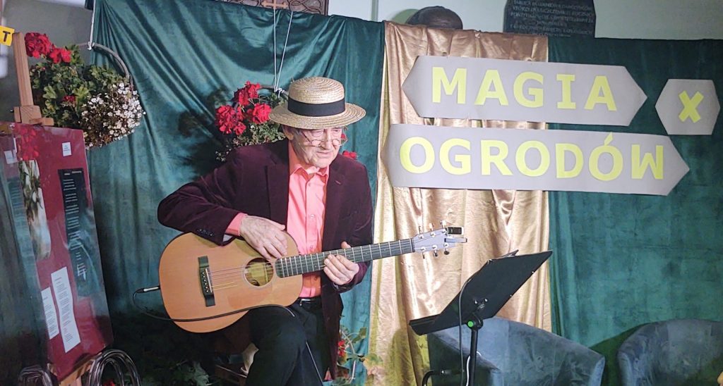 Aktor grający na gitarze na tle dekoracji z napisem Magia Ogrodów X 